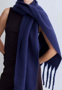 Marineblauwe wollen sjaal, zachte textuur, rechthoekige vorm, met franjes aan de uiteinden, over de schouder van een model gedrapeerd, gecombineerd met een zwarte top.