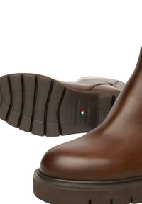 Bottes à cheville en cuir marron avec un bout arrondi, une semelle en caoutchouc épaisse avec des rainures, des accents cousus et un logo sur la semelle.