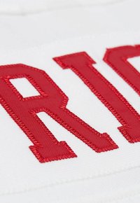 Nahaufnahme der rot gestickten Stoffbuchstaben "RIG" auf einem weißen strukturierten Stoffhintergrund.