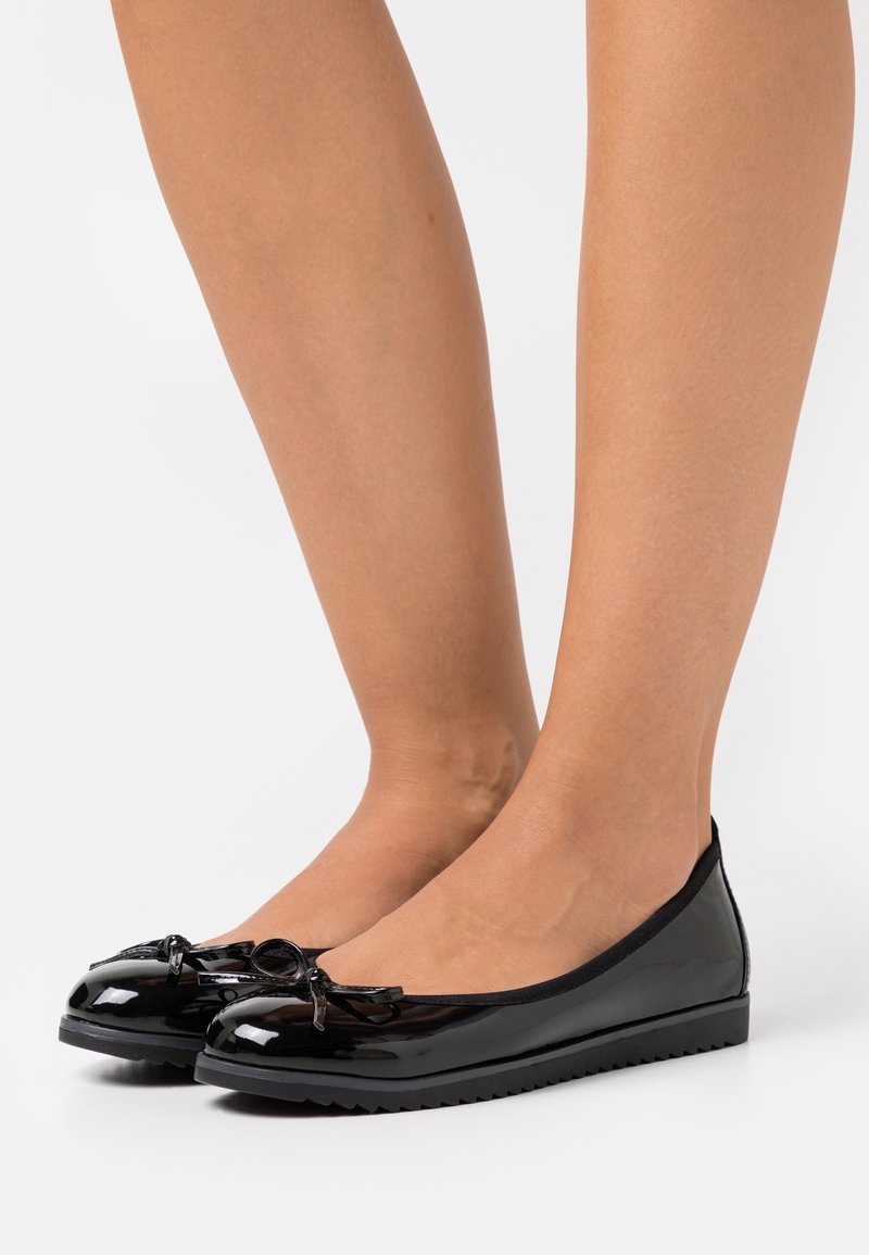 Ballerines en cuir verni noir brillantes avec petits nœuds, portées pieds nus sur un fond blanc uni.