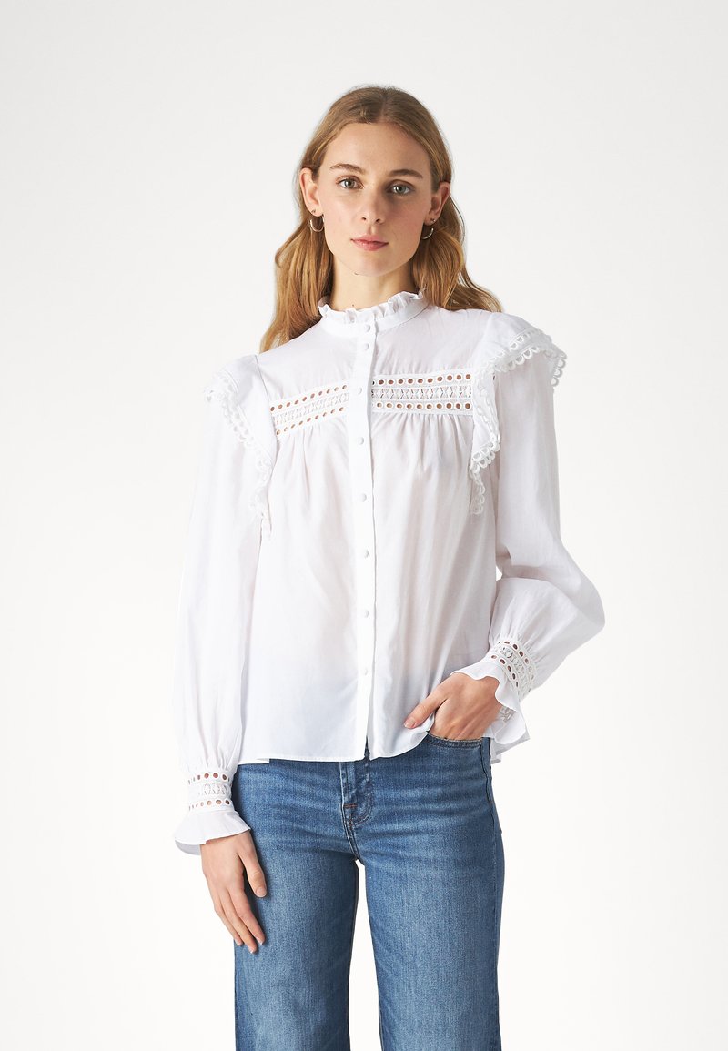 Vit bomullskjorta med volangärmar, spetsdetaljer och knäppning. Texturerad broderi och rynkning vid halsringningen. Kombinerad med blå jeans.