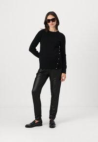 Svart genser med rund hals og knappdetaljer på siden, kombinert med tettsittende sorte, blanke bukser og svarte loafers. Ingen andre detaljer.