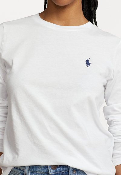 Langärmliges weißes Baumwoll-T-Shirt mit einem kleinen Navy-Logo auf der linken Brust, rundem Ausschnitt und lässiger Passform. Sichtbare blaue Jeans.