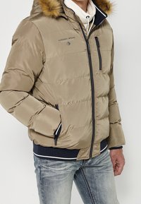 Homme portant une doudoune beige avec capuche bordée de fourrure, poche poitrine zippée et poignets côtelés foncés, assortie à un jean bleu clair délavé.