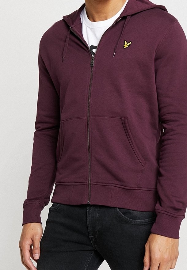 Homme portant un sweat à capuche bordeaux zippé avec un petit logo d'oiseau jaune sur la poitrine et un jean noir, debout devant un fond uni.