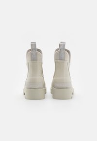 Botines beige con parte superior sintética lisa, cuello acolchado y detalles del logo en el talón y en la tira de agarre. La suela gruesa mejora la estabilidad.