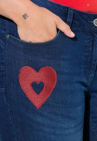 Džínové kalhoty s velkou červenou výšivkou srdce a menším výřezem ve tvaru srdce uprostřed, částečně viditelná přední kapsa.