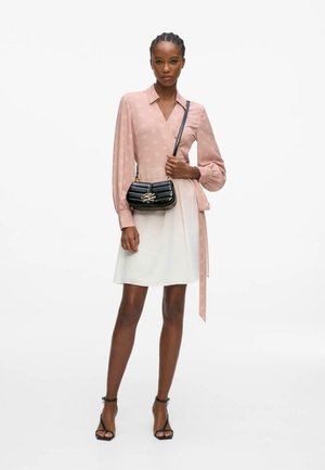 Mujer con blusa rosa de manga larga y falda blanca, sosteniendo un bolso negro acolchado de hombro con herrajes dorados, de pie con tacones negros.