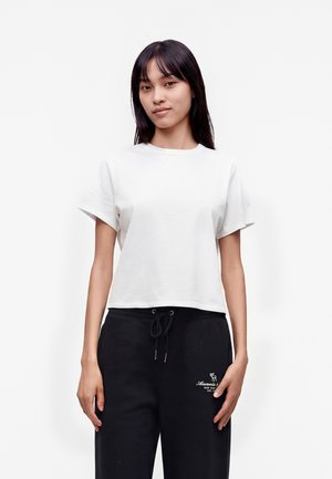 Vrouw met steil donker haar draagt een effen wit T-shirt met korte mouwen en zwarte broek met trekkoord, staand tegen een effen witte achtergrond.
