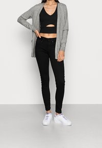 Grå lång cardigan över en svart crop top med ett utskärning, i kombination med svarta skinny jeans och vita sneakers, som skapar en lagerlook.