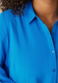 Lockigt hår över axeln, bärande en ljusblå knäppt skjorta med krage, som visar övre delen av bröstet och halslinjen.