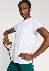 Weißes, kurzärmliges Sportshirt mit rundem Ausschnitt und strukturierten Einsätzen, kombiniert mit grünen Leggings. Das Modell hält Fitnessgeräte in der Hand.