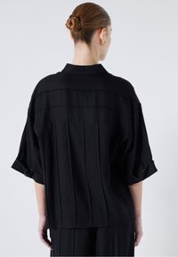 Chemise noire à coupe décontractée avec manches courtes, design plissé, texture douce et col. Vue de dos mettant en valeur des détails à rayures subtiles.