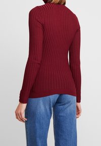 Burgundy ribbattu neule, jossa on korkea kaulus, pitkät hihat ja tiukka muoto. Kangas on pehmeä, hienovaraisella tekstuurilla ja tiukalla istuvuudella vyötäröllä.