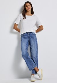 Kvinde med langt mørkt hår læner sig op ad en hvid væg, iført en hvid kortærmet skjorte, blå vide jeans og hvide sneakers.
