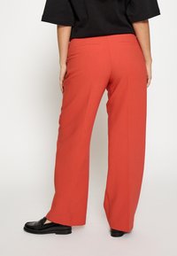 Rote breite Hosen aus glattem Stoff, mit zwei Gesäßtaschen und einer taillierten Passform, kombiniert mit schwarzen Slippern.