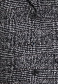 Manteau en laine à carreaux gris et noir avec trois boutons noirs en ligne verticale et une poche à rabat visible sur le côté droit.