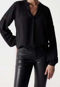 Blusa nera a maniche lunghe con scollo a V, caratterizzata da pieghe e dettagli in pizzo. Abbinata a pantaloni in pelle nera lucida, che enfatizzano un aspetto elegante e aderente.