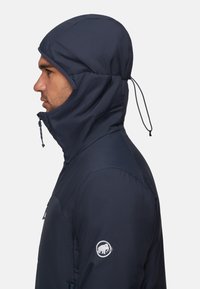 Uomo che indossa una giacca a vento blu navy con cappuccio e zip, con il logo Mammut sulla manica, rivolto a sinistra contro uno sfondo bianco uniforme.