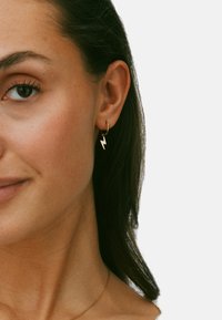 Zlatá kruhová náušnica so zúbkovan�ým tvarom. Náušnica visí na malom kruhu, ktorý dopĺňa prirodzený tvar ucha.