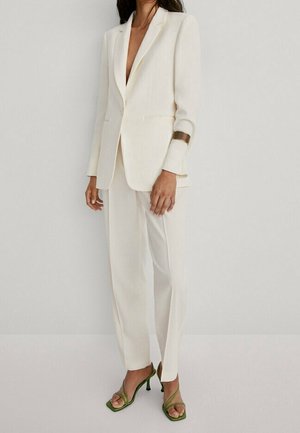 Frau trägt einen elfenbeinfarbenen, maßgeschneiderten Anzug mit einem Ein-Knopf-Blazer und geraden Hosen, kombiniert mit grünen, spaghetti-riemchen Sandaletten mit hohem Absatz.