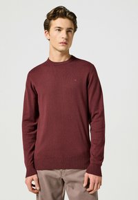 Maglione bordeaux a maniche lunghe in morbido materiale a maglia, con scollatura rotonda e polsini a costine. Piccolo logo sul petto.