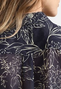 Marineblaue Bluse mit floralen Mustern in Creme. Leichter Stoff mit glatter Textur, dezentes Falten-Detail auf der Rückseite und klassischer Kragen.