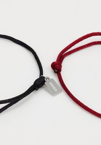 Bracelets en cordon tressé noir et rouge interconnectés par une petite étiquette en argent gravée du mot "HUGO" sur un fond blanc.