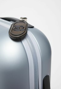 Gros plan sur un coin de valise rigide en argent avec une fermeture éclair et une étiquette de bagage noire attachée à la poignée.