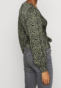 Blusa a fantasia leopardata in verde oliva, con maniche a sbuffo e vita annodata, abbinata a jeans grigi a vita alta. Tessuto morbido e testurizzato.