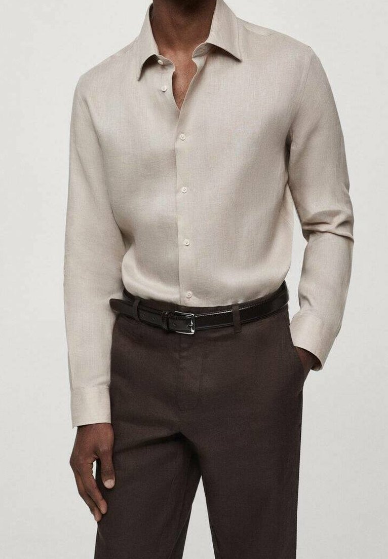Homme portant une chemise beige à manches longues boutonnée rentrée dans un pantalon marron foncé avec une ceinture noire, debout avec une main dans la poche.