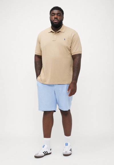 Polo beige avec un petit logo marine, shorts bleu clair et baskets blanches avec des rayures noires. Tissu lisse, coupe décontractée, shorts à mi-cuisse.