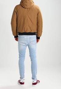 Brown padded jacket with a hood, black cuffs, paired with light blue fitted jeans and burgundy and white sneakers.