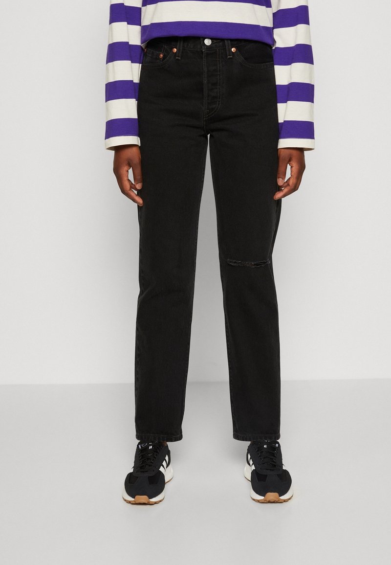 Personne portant un jean noir droit avec une petite déchirure au genou droit, des baskets noires et blanches, et un t-shirt à manches longues rayé violet et blanc.