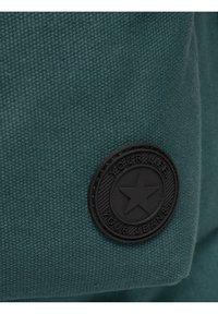 Žalias tekstilės pleistras su apvaliu juodu logotipu, kuriame yra iškilus žvaigždės dizainas ir išraižytas tekstas. Medžiaga turi austą tekstūrą.