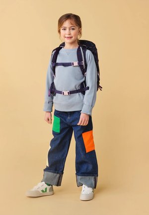 Felpa blu, pantaloni in denim con toppe arancioni e verdi, sneakers bianche con dettagli verdi e uno zaino viola con accessori metallici.