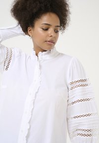 Chemise blanche à boutons avec des détails à volants le long de la patte de boutonnage et du col. Présente des accents en dentelle sur les manches avec une texture plissée.