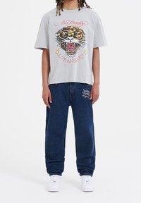 T-shirt grigio in cotone con una grafica colorata di una tigre e la scritta "Los Angeles", abbinato a pantaloni di jeans blu scuro e sneakers bianche.
