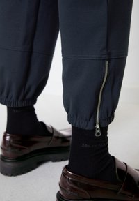 Tumši bikses ar elastīgām aprocēm un ar rāvējslēdzēju sānu kabatu, apvienotas ar tumšiem zeķēm un spīdīgiem brūniem loafers, kuriem ir biezas, teksturētas zolītes.