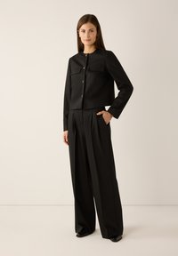 Black cropped jacket with two breast pockets and button closures paired with high-waisted black trousers; smooth fabric texture.