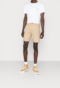 Hombre vistiendo una camiseta blanca de manga corta, pantalones cortos beige arremangados, calcetines blancos y zapatillas altas mostaza sobre un fondo liso.