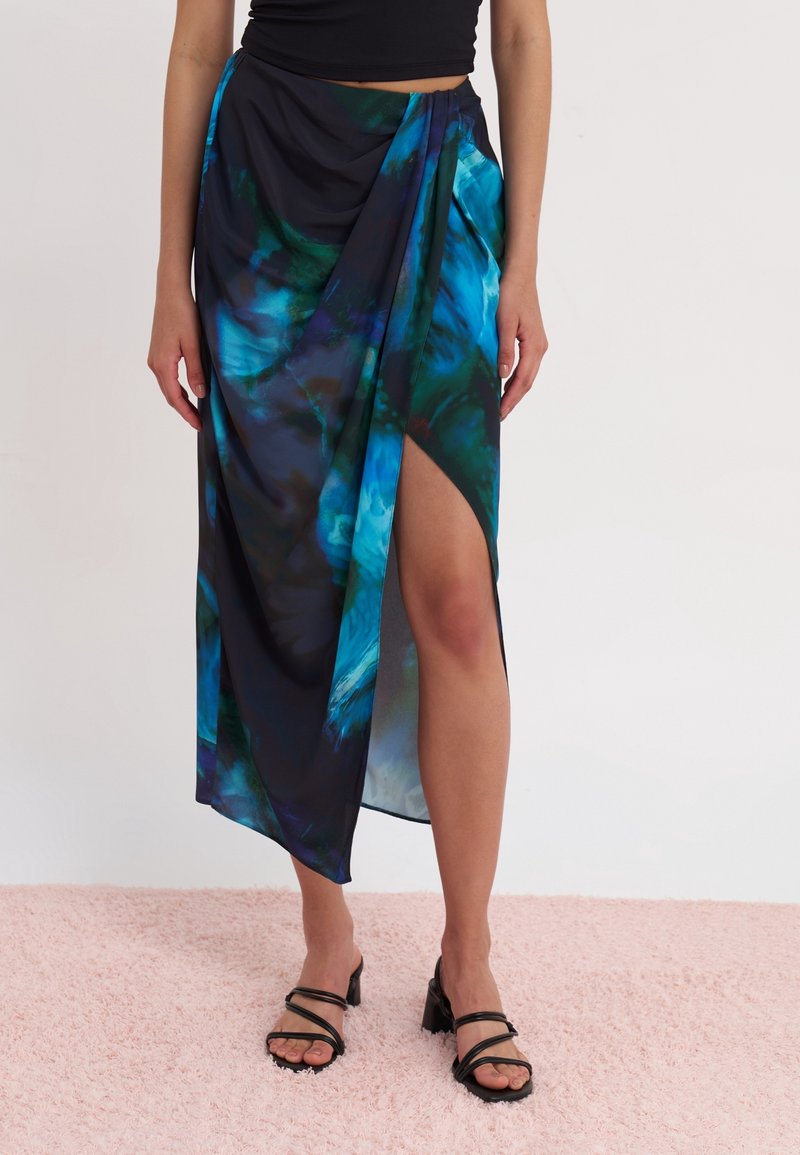 Femme portant une jupe portefeuille à imprimé abstrait bleu et noir avec une fente haute, associée à des sandales noires à talons et un top court noir.