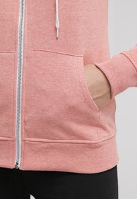 Persona vistiendo una sudadera con cremallera de color rosa claro, con una mano dentro del bolsillo frontal, combinada con pantalones negros.