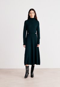 Knie-lang, dunkelgrünes Strickkleid mit langenÄrmeln und hohem Kragen. Verfügt über silberne Hardware-Akzente an der Taille und einen ausladenden Rock.