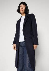 Marineblå uldfrakke med skræddersyet pasform, to knapper, klaplommer og let spidse revers, båret over en hvid top og blå jeans.