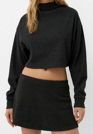 Femme portant un pull court noir côtelé à manches longues et une mini-jupe noire côtelée assortie laissant apparaître le ventre.