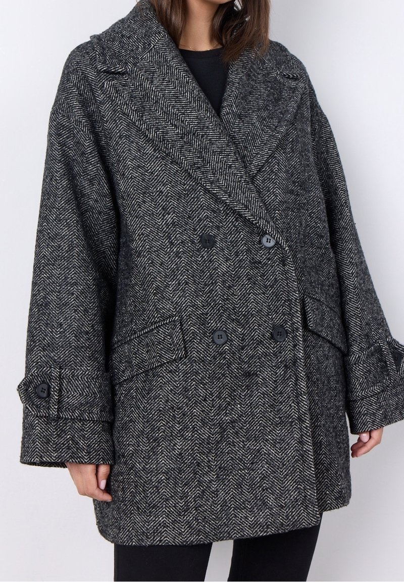 Płaszcz w wzór houndstooth z wełny, z podwójnym zapięciem, czarnymi guzikami i dużymi kieszeniami. Posiada szeroki kołnierz oraz luźne rękawy.