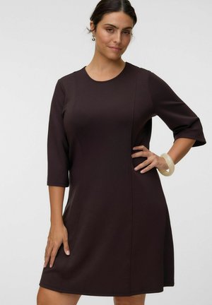 Femme portant une robe marron foncé, longueur genou, avec des manches trois-quarts, debout, une main sur la hanche, portant un bracelet clair.