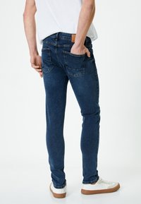 Jeans de mezclilla oscuros, ajustados, con un acabado ligeramente desgastado, que cuentan con dos bolsillos traseros y un parche de cuero marrón en la cinturilla.