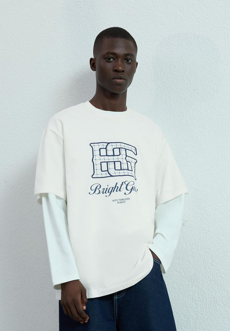 Biała, oversized koszulka z granatowym logo graficznym i napisem "Bright Gu" nałożonym na wzór siatki. Nosi się ją na jasnoniebieskiej, długiej koszulce.